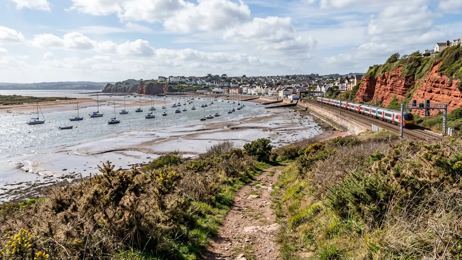 The Devon coast at half tide with moored boats on the sand and a railway line along the sea wall, rough salt bleached grass and scrub on the cliff bank in the foreground